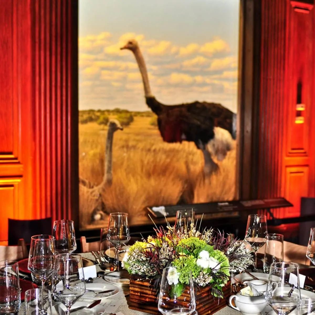 A dining room with a table set for a meal by a catering service in Los Angeles, overlooked by a large window showing an ostrich in a field at sunset.