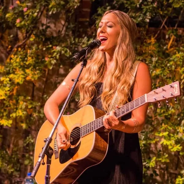 A woman singing and playing guitar onstage, surrounded by foliage, under warm catering lights in Los Angeles.